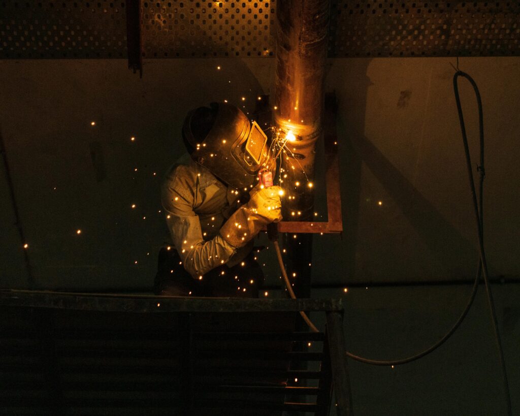 Pourquoi la maintenance préventive des pipelines est essentielle ? A welder at work indoors, creating bright sparks while welding metal pipes.
